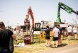 A group of people observe heavy machinery in action at an outdoor exhibition. The scene includes cranes, lifting equipment, and vehicles, with barriers placed around the demonstration area. One large orange crane holds a log vertically, while a green crane operates nearby. The event is taking place on a sunny day, with multiple visitors standing in the foreground, watching the machinery display.