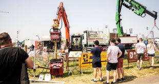 A group of people observe heavy machinery in action at an outdoor exhibition. The scene includes cranes, lifting equipment, and vehicles, with barriers placed around the demonstration area. One large orange crane holds a log vertically, while a green crane operates nearby. The event is taking place on a sunny day, with multiple visitors standing in the foreground, watching the machinery display.