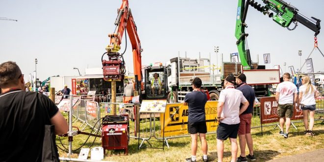 A group of people observe heavy machinery in action at an outdoor exhibition. The scene includes cranes, lifting equipment, and vehicles, with barriers placed around the demonstration area. One large orange crane holds a log vertically, while a green crane operates nearby. The event is taking place on a sunny day, with multiple visitors standing in the foreground, watching the machinery display.