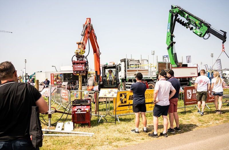 A group of people observe heavy machinery in action at an outdoor exhibition. The scene includes cranes, lifting equipment, and vehicles, with barriers placed around the demonstration area. One large orange crane holds a log vertically, while a green crane operates nearby. The event is taking place on a sunny day, with multiple visitors standing in the foreground, watching the machinery display.