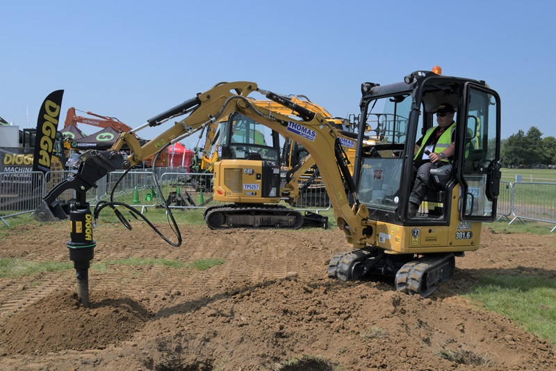  A yellow mini-excavator is in operation on a grassy site, fitted with a Digga attachment for drilling or digging. A construction worker, wearing a high-visibility vest, operates the machine. Behind the mini-excavator, another larger yellow excavator from the Thomas Plant fleet is visible. Both machines are at an outdoor event, surrounded by safety barriers and other construction equipment in the background.