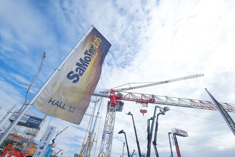 A flag reading "SaMoTer 24, Green Innovation" is seen fluttering at an outdoor construction machinery fair. The sky is partly cloudy, and cranes and other heavy equipment are visible in the background, indicating the event is focused on construction technology. The flag indicates the event is held in Hall 12.