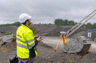 Construction worker operating a drone at a gravel quarry site with surveying equipment in the foreground.