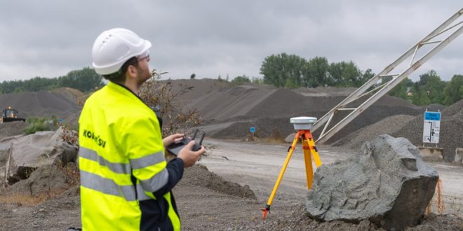 Construction worker operating a drone at a gravel quarry site with surveying equipment in the foreground.