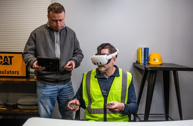 Worker using CAT simulator training with VR headset and controllers in a construction environment.