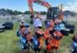 A group of young children wearing orange safety vests and caps pose with crossed arms in front of a large orange excavator at an outdoor event. An adult man and woman stand behind them making the same gesture. The children are holding colorful tote bags and appear to be enjoying the sunny day. Plantworx press release.