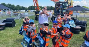 A group of young children wearing orange safety vests and caps pose with crossed arms in front of a large orange excavator at an outdoor event. An adult man and woman stand behind them making the same gesture. The children are holding colorful tote bags and appear to be enjoying the sunny day. Plantworx press release.