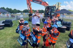 A group of young children wearing orange safety vests and caps pose with crossed arms in front of a large orange excavator at an outdoor event. An adult man and woman stand behind them making the same gesture. The children are holding colorful tote bags and appear to be enjoying the sunny day. Plantworx press release.