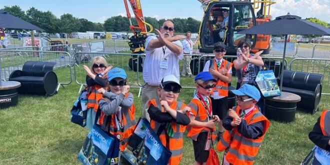A group of young children wearing orange safety vests and caps pose with crossed arms in front of a large orange excavator at an outdoor event. An adult man and woman stand behind them making the same gesture. The children are holding colorful tote bags and appear to be enjoying the sunny day. Plantworx press release.