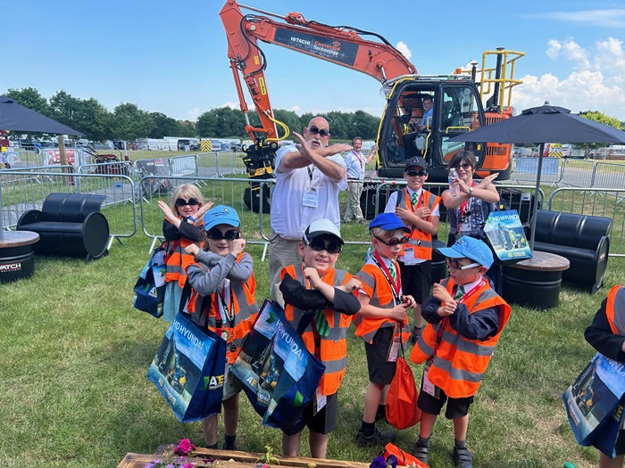 A group of young children wearing orange safety vests and caps pose with crossed arms in front of a large orange excavator at an outdoor event. An adult man and woman stand behind them making the same gesture. The children are holding colorful tote bags and appear to be enjoying the sunny day. Plantworx press release.