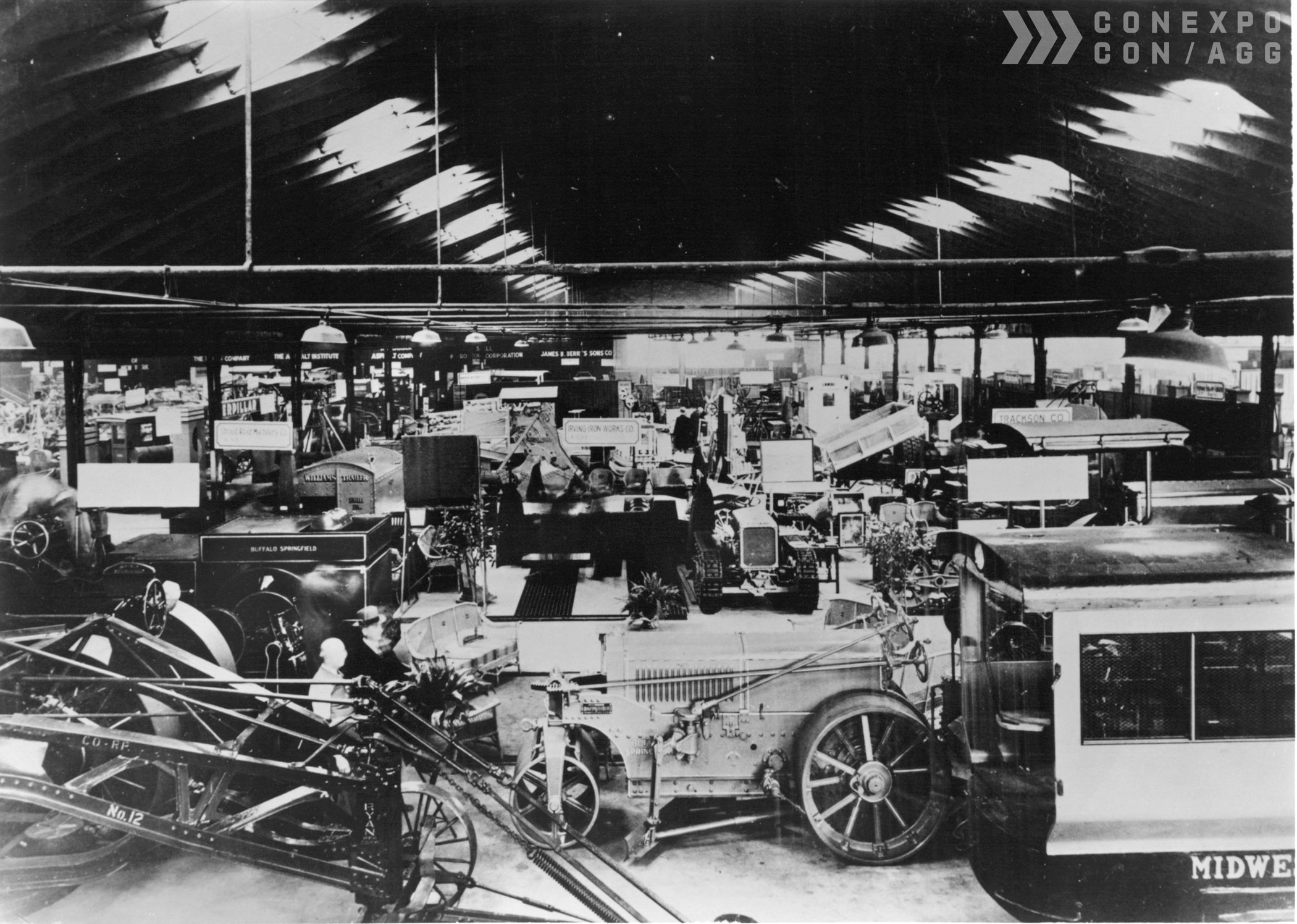 Historic indoor view of a mid-1900s construction equipment exhibition with cranes and large machinery.
